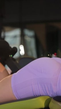Spor kıyafetli bir kadın spor salonundaki bir makineye bacak eklentisi yapıyor. Rahat etmesini ve kişisel koruyucu ekipman kullanmasını sağlıyor. Dikey video