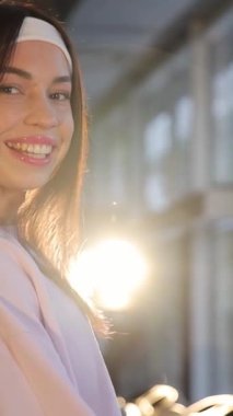 Saçları katmanlı ve saç bandı takmış sarışın bir kadın spor salonunun penceresinin önünde resmi bir şekilde gülümsüyor. Uzun kirpikleri var ve gözlük takıyor. Dikey video