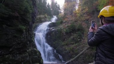 Huzurlu bir videoda bir kadın ve bir başkası çarpıcı bir şelaleden zevk alıyor, hevesle fotoğraf çekiyor, doğal huzuru somutlaştırıyor.