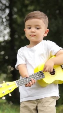 Genç müzisyen çocuk parktaki çimlerin üzerinde zevkle sarı ukulele çalıyor, sessiz bir çayırda güzel müzik yapıyor. Dikey video