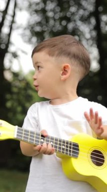 Bir çocuk müzisyen parktaki çimlerin üzerinde sarı ukulele çalmaktan ve parkta iyi müzik yapmaktan mutluluk duyar. Dikey video