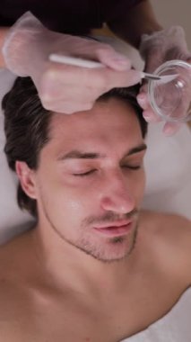 Bir adam kaplıcada yüz bakımı görüyor, burnuna, yanaklarına, gözlerine, kirpiklerine ve ağzına odaklanıyor. Terapist ayrıca kollarına masaj yapıyor ve yüz kaslarını gevşetiyor. Dikey video.