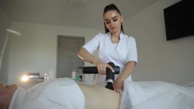 Genç kadın terapist, fizyoterapi sürecinde masaj masasında yatan kadın hastaya perküsyon silahı kullanıyor. Yüksek kalite 4k görüntü