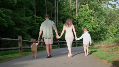Bir aile, neşeli köpekleriyle neşeyle el ele tutuşup, huzurlu bir ormanlık yolda yürür. Doğada beraberliği somutlaştırır.