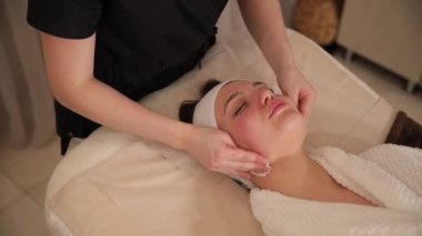 Lüks bir kaplıcada yüz masajı yaptıran bir kadının videosu. Estetisyen cildini tazelemek için teknikler kullanır, onu şımartır ve tazeler.