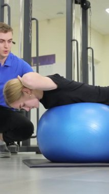 Bir erkek, spor salonunda eklem ve diz hareketlerine odaklanırken kollarındaki, bacaklarındaki ve kalçalarındaki kasları hedef alan pilates topu üzerinde egzersiz yapan bir kadına yardım ediyor. Dikey video