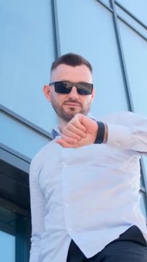 Uzun bir gökdelenin arka planında duran dikey videolu genç adam saate bakıyor. Şirketin klas çalışanı iş merkezinin arka planına karşı