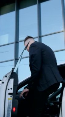 Takım elbiseli dikey video işadamı şehirdeki cam bir binanın yakınındaki beyaz bir arabayla yola çıkıyor. Şirketin başı. Zengin bir adam.