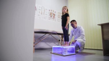 Klinikte, bir doktor genç bir kızın ayaklarını muayene eder, rahat etmesini sağlar. Bu, nazik çocuk bakımının önemini vurgular.