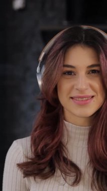 Amerikan görünümlü, hoş bir gülümsemesi olan pozitif bir kız dijital bir mağazada yeni kulaklık takıyor ve kameraya bakıyor. Yüksek kaliteli kulaklık sesi. Dikey video