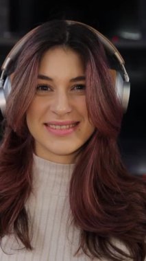 Amerikan görünümlü, hoş bir gülümsemesi olan dikey videolu kız dijital bir mağazada yeni kulaklık takıyor ve kameraya bakıyor. Yüksek kaliteli kulaklık sesi.