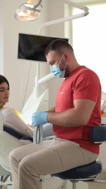 Tıbbi maskeli dikey video dişçisi bir hastayla tedavi planını tartışıyor. Dişçi randevusundaki genç bir kadın.