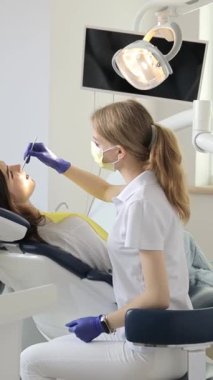 Diş tedavisi sırasında dişçi koltuğunda oturan dikey videolu kadın. Modern bir diş muayenehanesinde genç bir dişçi doktor ve hasta..