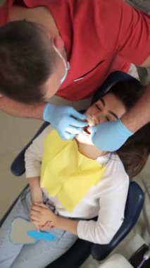 Dişçi koltuğundaki dikey videolu kadın. Bir dişçi, bir hastayı diş tedavisi için ağız koruyucusu takarak hazırlar. Ağız boşluğunda rahat bir tedavi..