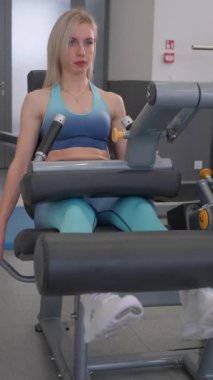 Dikey videoda bir kadın spor makineleri üzerinde bacak egzersizleri yapıyor ve güç eğitimini vurguluyor. Enerjik bir spor salonundaki bacak uzatma makinesinde uygun formu gösteriyor.
