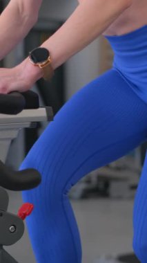 Dikey videoda bir kadın spor bisikleti sürüyor. Spor ve sağlık için pedal çevirirken kaslarını çalıştırıyor.