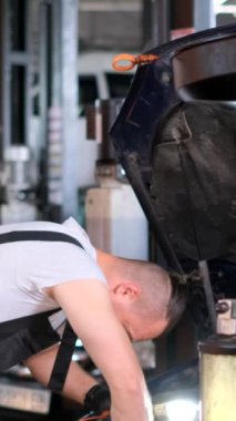 Dikey video. Bir tamirci, kaputu açık, otomobil parçaları ve aletlerle çevrili bir garajda bir motorlu araç üzerinde çalışıyor.