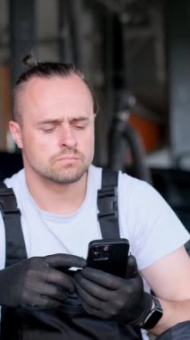 Sakallı bir müzisyen garajda yerde oturuyor, tişört ve yelek giyiyor. Otomotiv lastikleri ve tekerleklerle çevrili cep telefonuna bakıyor. Dikey video a