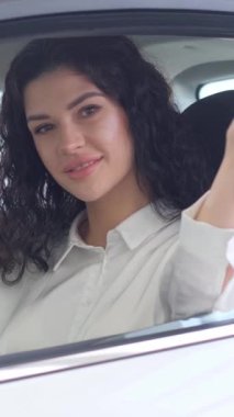 Dikey videoda bir kadın yeni bir araba aldı ve araba anahtarlarını gösterdi. Güzel saçlı çekici esmer kadın pahalı beyaz bir arabada oturur ve anahtarları gösterir.