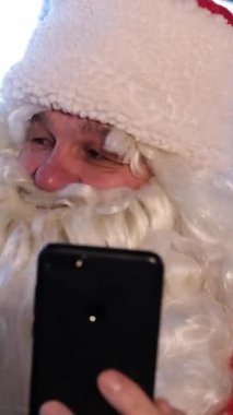 Şenlikli bir ortamda, Noel Baba sevinçle selfie çeker ve güzel dekore edilmiş bir Noel ağacının yanında neşeli kişiliğini gösterir.