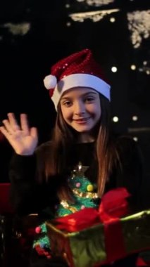 İç açıcı bir tatil videosunda Noel Baba şapkalı bir kız Noel ağacının yanında kamerayla neşe içinde el sallıyor ve öpücükler savuruyor.