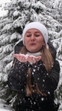 Bir kadın, canlı beyaz kar taneleriyle dolu güzel bir karlı ormanın önünde ellerinden neşeyle kar üflüyor.