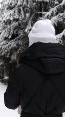 Siyah ceketli ve beyaz şapkalı bir kadın kar kaplı bir arazide yürüyor, sakin kış manzarasının tadını çıkarmak için mola veriyor.