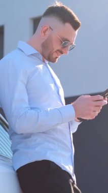 Güneş gözlüklü ince bir işadamı pahalı arabanın yanındaki telefona bakıyor. Güneşli bir günde sokakta meşgul bir işadamı. Dikey video