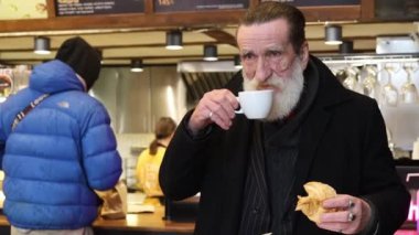 Sıcak bir kafede, yaşlı bir adam sabah rutininin tadını çıkarıyor, kahvesini yudumluyor ve sıcak bir atmosferde lezzetli bir sandviçin tadını çıkarıyor.