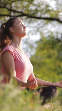 Nilüfer pozisyonunda oturan bir kadın huzur parkında çimlerin üzerinde, kapalı gözlerle meditasyon yapıyor, güzellikle çevrili, davetkar bir sakinlik.