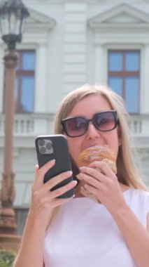 Dikey videolu genç kız, güzel bir binanın arka planında dikilirken telefona bakarken dondurma yiyor. Güneş gözlüklü bir kız yaz günü yürür ve dondurma yer.