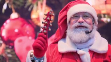 Pozitif Noel Baba, izleyicilerle mikrofona konuşuyor, parlak bir Noel arka planında duruyor, değneğini sallıyor. Yeni yıl Noel Baba 'sı insanlara mutlu yıllar ve Noel diliyor.