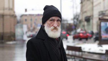 Sakallı ve siyah şapkalı bir adam işlek bir şehir caddesinde durmuş, etrafındakileri merakla izliyor.