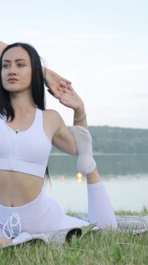 Dikey video esmer, yoga dersleri sırasında sabah gölünün arka planına doğru esneme hareketleri yapıyor. Göl kenarında açık hava yogası.