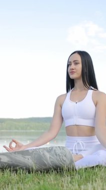 Dikey video, güzel esmer kadın sabah gölünde yoga yaparken lotus pozisyonunda oturuyor. Göl kenarındaki açık havada yoga..