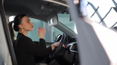 Yeni arabasında bir araba galerisinde oturan ve aynaya bakan genç, gülümseyen bir iş kadını. Yüksek kalite 4k görüntü
