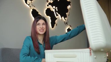 Bir kadın arka planda dünya haritası olan bir bavulu açıyor, haritanın önünde beyaz bir kutu tutuyor, keşif ve macera ruhunu çağrıştırıyor.
