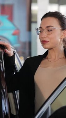 Dikey video, araba galerisinde açık bir arabaya yaslanan genç bir iş kadını, araba galerisi. İş kadını yeni bir araba seçer.