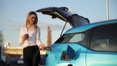 Bir kadın telefonunu kullanırken bir istasyonda elektrikli arabasını şarj ediyor. Çevre dostu ulaşım kavramını gösteriyor.