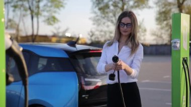 Elektrikli benzin istasyonunda sarı saçlı genç bir kadın elektro arabayı şarj ediyor. Yüksek kalite 4k görüntü