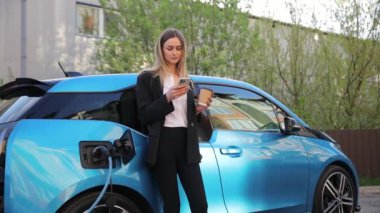 Elektrikli bir arabanın yanında durup şarj olurken telefonunu izleyen bir kadın. Mobil cihazıyla birden fazla iş yaparak şarj sürecine odaklanmış durumda.