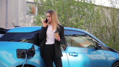 Takım elbiseli güzel sarışın kadın telefonda mavi şarjlı bir arabanın yanında konuşuyor. Yüksek kalite 4k görüntü