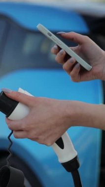 Dikey video, kadın elektrikli arabasının yanında duruyor, cep telefonunu şarj ediyor. Araç kaputu açık, lastik ve tekerlek gibi otomotiv parçalarını gösteriyor.