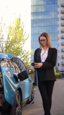 İş elbisesi giymiş profesyonel bir kadın elektrikli arabasından para alıyor. Mavi bir elektrikli arabanın yanında duruyor. Çevresel ulaşımın ve modern teknolojinin altını çiziyor.