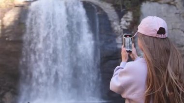 Resimli bir yerde duran bir kadın, hatıralar için cep telefonuyla çarpıcı bir şelalenin güzel bir fotoğrafını çekiyor.