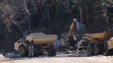 Video bir inşaat sahasında ağır makineler gösteriyor, buldozer ve çöp kamyonu taşları hareket ettiriyor, ekipman koordinasyonunu belirliyor.