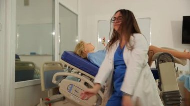 Tıbbi kıyafetli bir kız, jinekoloji koltuğunda hamile bir kadının arka planında dans ediyor.