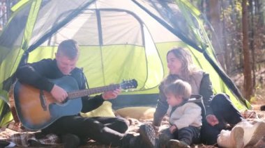 Bir adam, aile ve iki çocukla çevrili rahat bir çadırın önünde gitar çalıyor, neşeli anılar yaratıyor ve beraberliği vurguluyor.