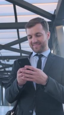 Resmi takım elbise giymiş ve kravat takmış bir adam cep telefonuna dikkatle bakıyor, ekranda önemli bir şeye odaklanmış gibi görünüyor.