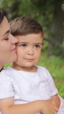 Dikey videoda anne ve baba küçük oğullarını öperler, yeşil ağaçların arka planında. Mutlu aile kavramı. Yüksek kalite 4k görüntü